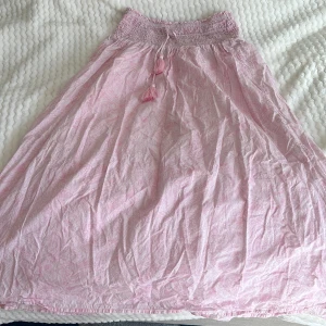 Rosa långkjol med smock och tofsar - Säljer en söt rosa långkjol med smock i midjan och dekorativa tofsar. Kjolen har ett diskret mönster och är perfekt för varma dagar. Den är luftig och bekväm att bära.❤️