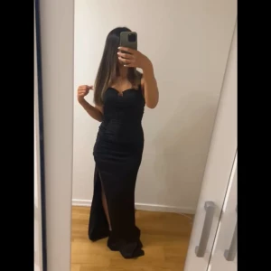 Svart långklänning med slits - Elegant svart långklänning med smickrande passform och slits framtill. Klänningen har smala axelband och en stilren design som passar perfekt för festliga tillfällen.