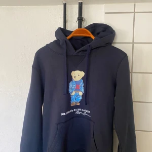 Mörkblå hoodie från Polo Ralph Lauren - Säljer en snygg mörkblå hoodie från Polo Ralph Lauren med den ikoniska Polo Bear på framsidan. Tröjan har en klassisk huva med dragsko och en stor magficka. Perfekt för en avslappnad stil! Säljer för att den är lite för liten. (Är 180 cm och runt 76 kg). Pris kan diskuteras!!