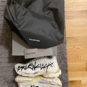 Vita sneakers från Balenciaga - Snygga vita sneakers från Balenciaga med svart graffiti-liknande text på sidorna. Skorna har en chunky sula och snörning framtill. Perfekta för dig som vill ha en unik och trendig stil.