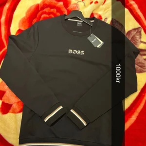 Svart tröja från Boss - Äkta!!! Snygg svart tröja från Boss med märkets logga broderad på bröstet. Tröjan har långa ärmar och ribbade muddar med vita och beige detaljer. Perfekt för en stilren look.