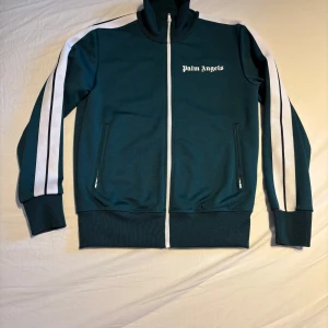 Grön track jacket från Palm Angels - Snygg grön track jacket från Palm Angels med vita ränder längs ärmarna och dragkedja framtill. Jackan har en hög krage och två fickor med dragkedja. Perfekt som tränings tröja eller bara en exklusiv look. Tröjan är använd ca 5 gånger så med andra ord nästan ny. 
