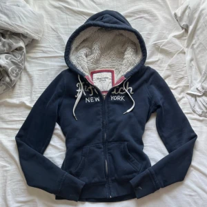 Mörkblå hoodie Abercrombie & Fitch - Mysig mörkblå hoodie från Abercrombie & Fitch med broderad logga och text på bröstet. Fodrad med fluffigt vitt teddyfoder, har huva med snörning och dragkedja framtill. Perfekt för kalla dagar och chill vibes. Sitter som xs💕