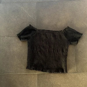 Svart smockad topp från Primark - Svart smockad topp från Primark med korta ärmar och offshoulder-modell. Toppens material är stretchigt och har en rynkad struktur som ger en cool och trendig look. Perfekt att matcha med höga jeans eller kjol för en chill stil.