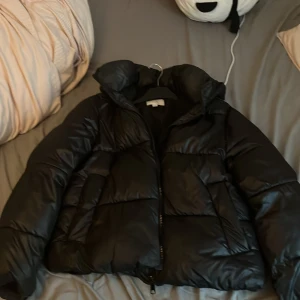 Svart pufferjacka från H&M, M - Säljer en svart pufferjacka från H&M i storlek M. Jackan har en hög krage, huva och dragkedja framtill. Den är vadderad och har en oversized look, perfekt för kalla dagar. Enkel och stilren design som funkar till många outfits.