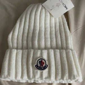 Vit stickad mössa från Moncler - Vit ribbstickad mössa från Moncler med uppvikt kant och klassisk logotyp framtill. Tillverkad i mjukt stickat material som håller dig varm och snygg under kalla dagar. Perfekt accessoar för vinterlooken. FINNS FLER MODELLER