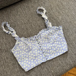 Blommig blå linnetopp från Zara - Supersöt croppad linnetopp från Zara i ljusblått med små vita och ljusgröna blommor. Toppen har volangdetaljer längs axelband och kant, knappar framtill och smockad rygg för extra komfort. Perfekt för varma dagar och en chill sommarstil.