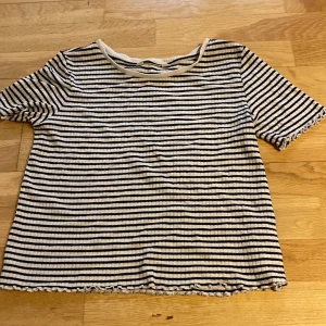 Randig ribbad topp från H&M - Säljer en svartvit randig topp från H&M i ribbad bomull. Modellen har korta ärmar och vågig kant nertill och vid ärmslut. Perfekt för en chill och stilren look.