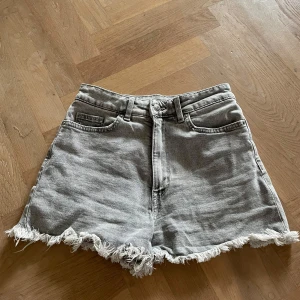 Grå jeansshorts med fransig kant - Snygga grå jeansshorts med hög midja och rå, fransig kant nertill. Klassisk femficksmodell med bälteshällor och dragkedja fram. Perfekta för sommardagar när du vill ha en avslappnad och trendig look.