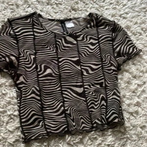 Brun zebramönstrad mesh topp från Divided XS - Säljer en kortärmad, croppad mesh topp från Divided i storlek XS. Toppen har ett brunt och beige zebramönster och är tillverkad i ett tunt, transparent syntetmaterial. Jättegulig, använts några gånger, pris går att diskuteras