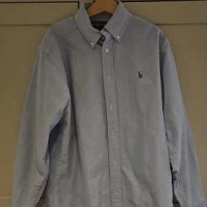 Ljusblå skjorta från Ralph Lauren - Klassisk ljusblå skjorta från Ralph Lauren med button-down krage och broderad logga på bröstet. Skjortan är tillverkad i 100% bomull och har långa ärmar samt knäppning framtill. Perfekt för en stilren och avslappnad look.