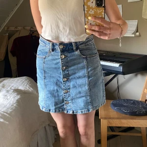 Blå jeanskjol från Mango - Snygg blå jeanskjol i modellen Vicky från Mango med uppknäppningsbara knappar hela vägen och bälteshälllor. Perfekt att styla med en crop top eller hoodie för en chill look.