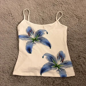 Vit blommig linnetopp från Bershka - Supersöt vit linnetopp från Bershka med tunna axelband och två stora blå liljor framtill. Har aldrig använts! Toppen är figurnära och har en fräsch sommarkänsla, perfekt till jeans eller kjol. Materialet är mjukt och stretchigt, vilket gör den riktigt bekväm.