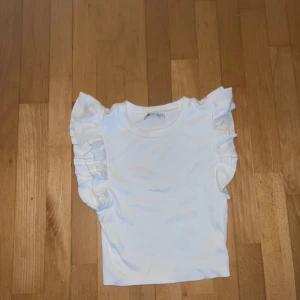 Vit volangtopp med ärmlös design - Supersöt vit topp med volangdetaljer på axlarna som ger en extra touch. Ärmlös modell med rund halsringning och croppad passform. Den är lite liten i storleken. Perfekt för dig som vill ha en enkel men ändå trendig look.