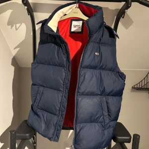 Mörkblå dunväst från Tommy Jeans - Snygg mörkblå dunväst från Tommy Jeans med röd insida och huva. Västen har quiltad design, dragkedja framtill och två stora fickor. Perfekt för lager-på-lager och ger en sportig vibe. Klassisk logga på bröstet och cool kontrast mellan färgerna.