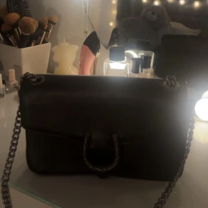 Svart axelväska med kedjerem - Super snygg svart axelväska i skinn med silverfärgad kedjerem och spänne framtill. Snygg och smidig. Säljer då den tyvärr inte kommer till användning längre.