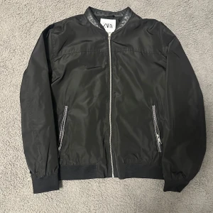 Svart bomberjacka från Zara - Svart bomberjacka från Zara med klassisk ribbad mudd i ärmslut och nederkant. Jackan har dragkedja framtill, två snedställda fickor med dragkedja och en stilren, minimalistisk look. Materialet är blankt och lätt, perfekt för en clean streetstyle.