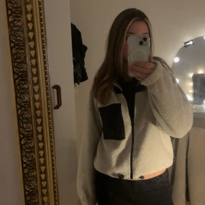 Beige teddyjacka med svart ficka - Mysig beige/krämvit teddyjacka med svart dragkedja och svarta detaljer. Går att spänna nere vid midjan, och har även en ficka. Köptes här på plick men är ifrån Gina Tricot.