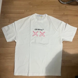 Vit Off-White t-shirt med tryck - Vit t-shirt från Off-White med svart och rosa tryck framtill och stort grafiskt tryck på ryggen med text, pilar och illustration. Oversized passform och klassisk rund hals. Materialet är mjuk bomull, perfekt för en chill och trendig look.