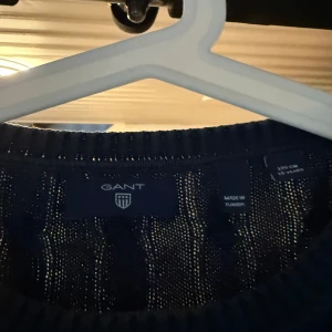Mörkblå kabelstickad tröja GANT - Snygg mörkblå kabelstickad tröja från GANT med rund halsringning och diskret logga på bröstet. Tröjan har långa ärmar och ribbade muddar vid ärmslut och nederkant. Perfekt för dig som gillar klassisk och stilren look. Lite blekt från tvätt men inget som syns 