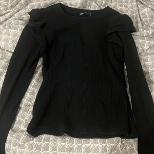 Svart ribbad topp med puffärm från Zara - Svart långärmad topp från Zara med ribbad struktur och cool puffdetalj på axlarna. Sitter snyggt med en lite tightare passform och rund halsringning. Perfekt för dig som gillar stilrena plagg med en twist.