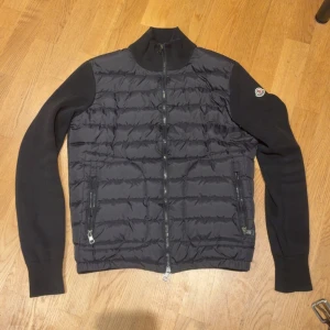 Svart Moncler cardigan med stickade ärmar - Snygg svart pufferjacka från Moncler med quiltad framsida och stickade ärmar. Jackan har hög krage, två dragkedjefickor och Moncler-logga på ärmen. Perfekt för kyliga dagar när du vill ha både stil och komfort. Fint skick. Har moncler mail äkthetsbevis.