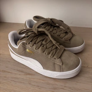 Puma Suede XL sneakers brun/vit - Snygga Puma Suede XL sneakers i brun mocka med breda snören och vit sula. Klassisk design med vit sidodetalj och guldfärgad Puma-logga på sidan. Skorna har en låg profil och är perfekta för en clean streetstil.