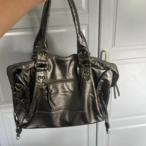 Metallic axelväska med detaljer - Snygg metallicfärgad axelväska i syntet med mässingsfärgade metalldetaljer och dragkedjor. Väskan har både handtag och avtagbar axelrem, samt flera fack med dragkedja framtill. Perfekt storlek för allt du behöver ha med dig och cool design.