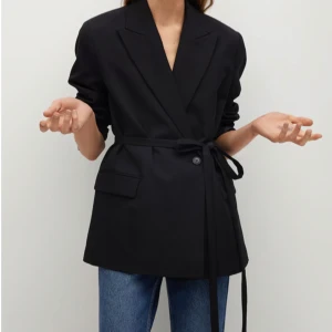 Svart kavaj från MANGO med bälte - Stilren Belt sugit blazer från MNG i storlek 38. Klassisk design med slag, två fickor framtill och tillhörande bälte för att knyta i midjan. Perfekt för en snygg och uppklädd look. Materialet känns mjukt och följsamt.