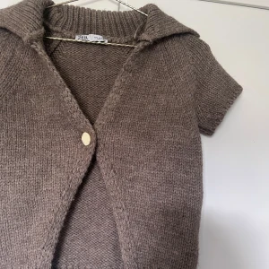 Beige stickad kofta från Zara - Kortärmad stickad kofta från Zara i en snygg beige nyans. Koftan har en stor krage, en enkel vit knapp framtill och är gjord i ett mjukt, grovstickat material. Perfekt att slänga över ett linne eller t-shirt för en chill look.
