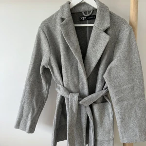 Grå kappa med bälte från Zara - Snygg grå kappa från Zara i klassisk modell med bälte i midjan och stora slag. Jackan har långa ärmar och två stora fickor framtill. 