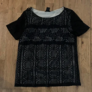 Svart spetsblus med mönster från Lindex - Supersnygg svart blus från Lindex i storlek XS. Blusen har korta ärmar och är täckt av ett detaljerat spetsmönster med geometriska former. Den är fodrad med ljus insida och har en klassisk rund halsringning. Perfekt för dig som gillar unika detaljer!