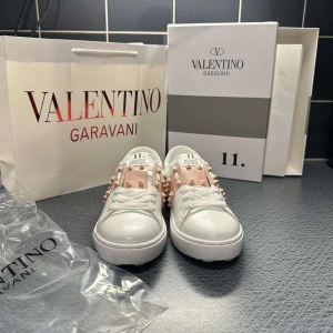 Valentino Rockstud sneakers vit/rosa - Snygga Valentino Rockstud sneakers i vitt skinn med ljusrosa band och ikoniska nitar. Klassisk rund tå, platt sula och coola detaljer som gör dem till ett statement. Perfekta för dig som vill sticka ut med exklusiv streetstyle.