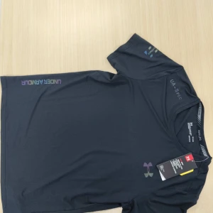 Svart t-shirt från Under Armour - Svart t-shirt från Under Armour med diskret logga på bröstet och tryck på axeln. Gjord i ett lätt och snabbtorkande material som passar perfekt till träning. Klassisk rund hals och korta ärmar. Snygg och sportig vibe.