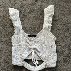 Vit broderad topp med volang från Bik Bok - Superfin vit topp från Bik Bok med broderade detaljer och volangiga axelband. Toppen är croppad, har knyt framtill och är ärmlös. Perfekt för sommardagar och ger en romantisk vibe med sin spets och luftiga känsla.