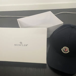Svart keps från Moncler - Snygg svart keps från Moncler med klassisk böjd skärm och broderad logotyp framtill. Kepsen har en stilren design och är tillverkad i ett mjukt material som sitter bekvämt på huvudet. Perfekt accessoar för en clean och sportig look.