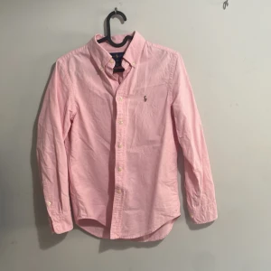 Rosa skjorta från Ralph Lauren - Snygg rosa skjorta från Ralph Lauren med klassisk button-down krage och broderad logga på bröstet. Skjortan har långa ärmar, knappar framtill och är tillverkad i mjuk bomull. Perfekt för dig som gillar stilrena och tidlösa plagg. Sitter som xs 