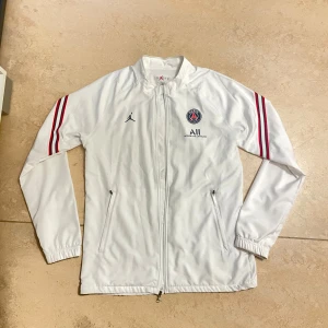 Vit PSG x Jordan vindjacka M - Snygg vit PSG x Jordan vindjacka med röda och marinblå ränder på ärmarna. Jackan har dragkedja framtill, två fickor med dragkedja och elastiska muddar. Perfekt för dig som gillar sportig stil och vill sticka ut med en ikonisk look. Priset kan också diskuteras vid snabb affär.