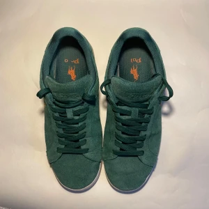Gröna Skor från Polo Ralph Lauren - Säljer ett par gröna sneakers i mocka från Polo Ralph Lauren med klassisk orange logga broderad på sidan. Skorna har vita sulor, rund tå och gröna skosnören. Perfekta för dig som gillar stilrena och sportiga skor med premiumkänsla! Skorna är i fint skick inga stora skador på mockan eller sulan. Tveka inte på att ställa frågor!🙌 Priset är inte hugget i sten! 🪨 