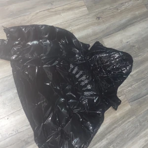 Svart pufferjacka från Trapstar - Säljer en svart, glansig pufferjacka från Trapstar med stor huva och fet logga broderad i vitt på ryggen. Jackan har bra kvalité och är knappt använd.