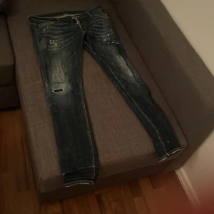 Dsquared2 blå slitna jeans - Säljer ett par blå jeans från Dsquared2 med coola slitningar och distressed detaljer. Jeansen har smal passform, klassisk femficksdesign och tydliga kontrastsömmar. Röd Dsquared2-patch bak i midjan och logotyp vid gylfen. Perfekta för dig som gillar streetstyle.