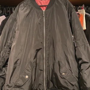 Svart bomberjacka Nirvana Divided XL - Svart bomberjacka från Divided i storlek XL med Nirvana-tryck på ryggen i rosa, blått och gult. Jackan har dragkedja framtill, snedställda fickor med tryckknappar och ribbade muddar. Fodret är rosa och materialet är glansig polyester.