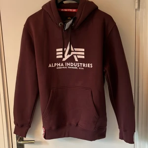 Vinröd hoodie Alpha Industries - Säljer en vinröd hoodie från Alpha Industries med vit logga och text framtill. Tröjan har huva med snörning, stor magficka och ribbade muddar. Perfekt för dig som gillar streetwear och vill ha en snygg och bekväm look.