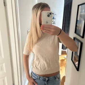 Beige kortärmad tröja från H&M - En stilren beige tröja från H&M med korta ärmar och rund hals. Tröjan är i mjuk bomullsmix och har ribbade detaljer vid hals, ärmslut och nederkant. Perfekt till jeans eller kjol för en clean look.