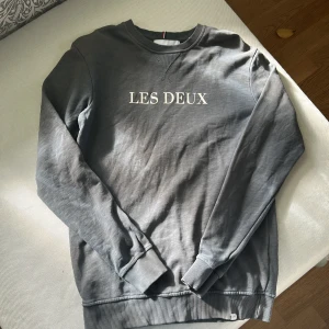 Grå sweatshirt från Les Deux - Säljer en stilren grå sweatshirt från Les Deux med vit text på bröstet. Tröjan har rund halsringning, ribbade muddar och är långärmad. Perfekt för en clean och avslappnad look. Materialet är mjuk bomull som känns skönt mot huden.