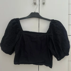 Svart croppad blus med puffärmar - Supersöt svart croppad blus med fyrkantig ringning och stora puffärmar. Ärmarna har broderade detaljer och resår vid ärmslut. Smockad rygg för extra bekväm passform. Perfekt till jeans eller kjol för en trendig look.