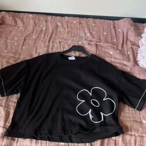 Svart croppad t-shirt från zara - Svart croppad t-shirt från Zara med en stor vit blomma tryckt framtill. T-shirten har korta ärmar och avslappnad passform. Perfekt för dig som gillar en chill och trendig stil.🩷