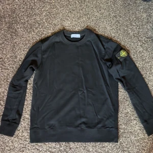 Svart Stone Island sweatshirt - Svart sweatshirt från Stone Island med klassisk rund hals och lång ärm. Tröjan har den ikoniska loggan på vänster ärm och är tillverkad i mjuk bomull. Enkel och stilren design som passar perfekt till jeans eller joggers.