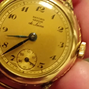 Guldfärgad vintageklocka Record Genève - Säljer en klassisk guldfärgad armbandsklocka från Record Genève, de Luxe. Klockan har rund boett med snygga detaljerade mönster på sidorna och en urtavla med svarta siffror och blå visare. Baksidan är graverad med 'Adina 1950'. Perfekt för dig som gillar retrostil.Nytt batteri behövs.