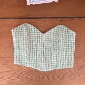Grön och vit rutig korsett från Zara - Superfin korsett från Zara i grönt och vitt rutigt mönster. Axelbandslös bandeau-modell med markerad sweetheart-urringning och stadig passform. Yttermaterialet är strukturerat och fodret är i slätt tyg. Perfekt att styla med jeans eller kjol för en trendig look.